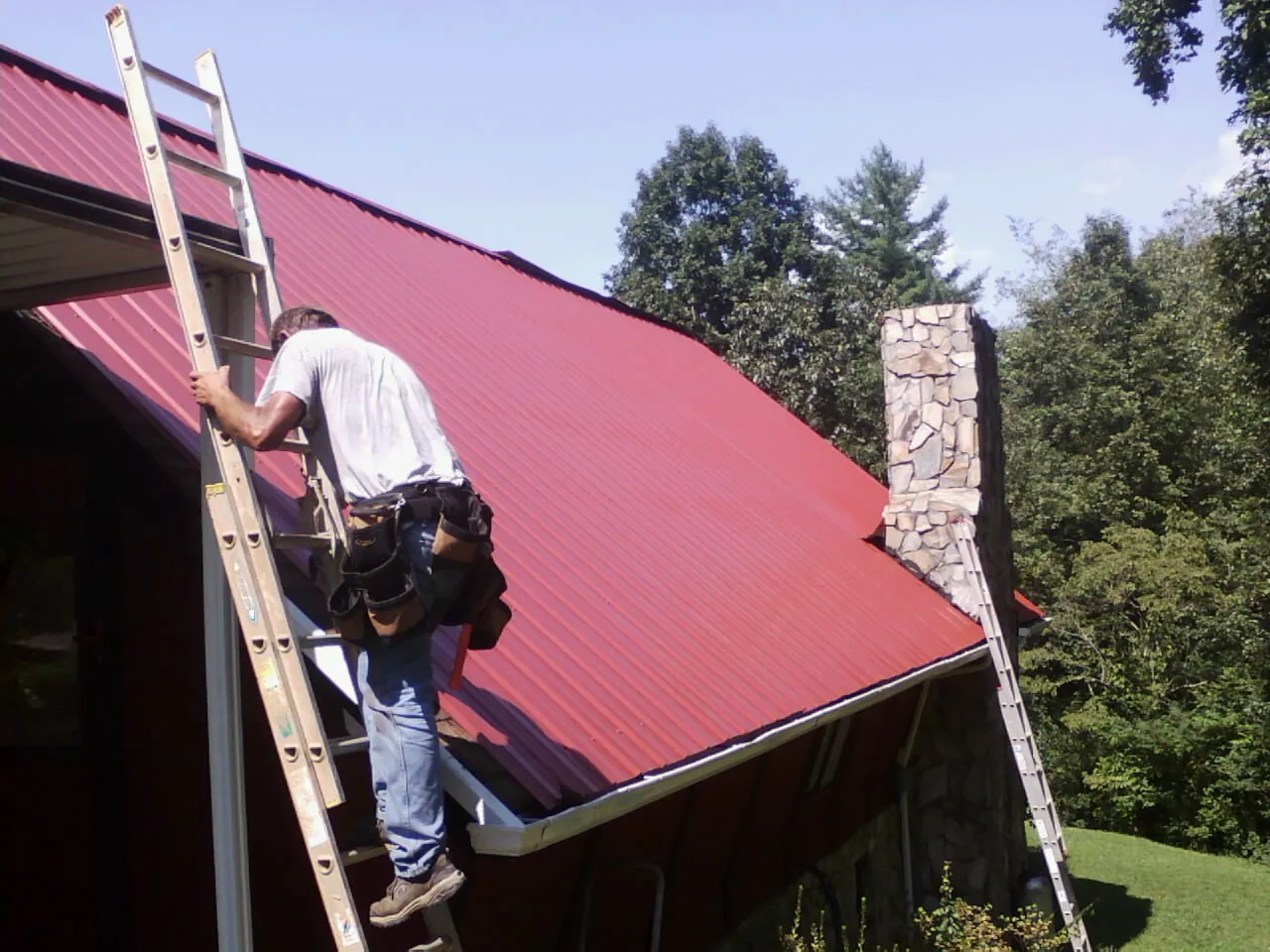 Roofer on a ladder beside a red metal roof and stone chimney — Soffit & Fascia Repair by Roofing Brothers in Euless